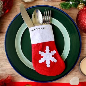 May include: A red and white stocking with the name "HARPER" spelled out in black letters. The stocking is filled with a fork, knife, and spoon. The stocking is sitting on a green plate with a white rim.