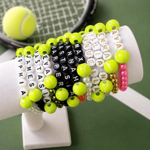 May include: A collection of beaded bracelets featuring white and black letter beads spelling names like Sophia, James, Eva, Max, Hannah, and Emma. The bracelets are accented with bright neon yellow tennis ball beads and some metallic gold beads.