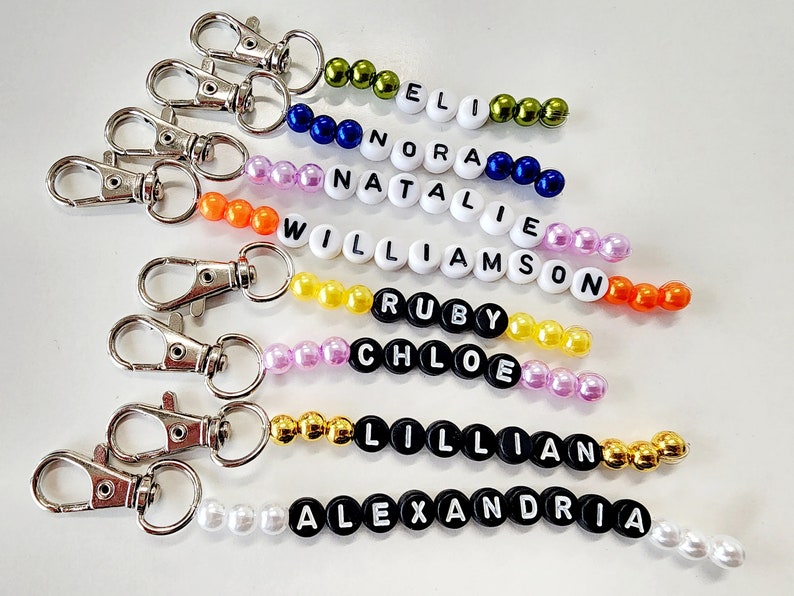 May include: A set of eight beaded keychains with different names spelled out in black letters. The beads are various colors, including green, blue, orange, yellow, pink, and white.
