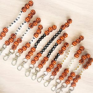 May include: A set of 10 basketball-themed keychain charms with personalized names. Each charm features a string of orange and black basketball beads with white letter beads spelling out a name. The charms are attached to silver key rings.