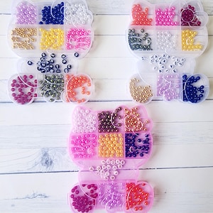 May include: Three pink plastic containers shaped like a cat with compartments filled with colorful beads. Each container has a row of letter beads at the bottom. The letters spell out "A-S-H-L-E-Y", "J-A-Z-M-I-N-E", and "G-A-B-R-I-E-L-L-E".