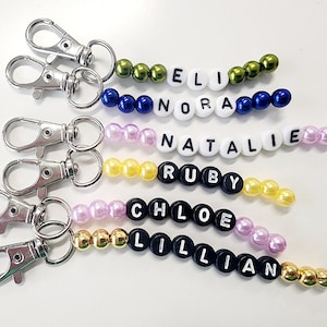 May include: Five colorful beaded keychains with different names spelled out on them. The names are Eli, Nora, Natalie, Ruby, and Lillian. Each keychain has a silver metal clasp.