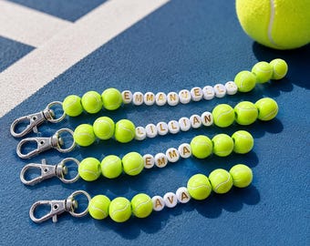 May include: Four keychains featuring lime green tennis ball beads and white letter beads spelling the names EMMANUELLA, LILLIAN, EMMA, and AVA. Each keychain includes a silver-toned metal lobster clasp for attachment to bags or other items.