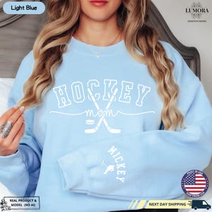 以下が含まれることがあります： 薄い青色のスウェットシャツに、白い文字で「HOCKEY MOM」と書いてあり、交差したホッケーのスティックとパックがあります。袖には「MICKEY」という文字があります。