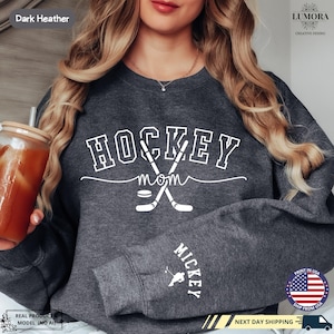 以下が含まれることがあります： ダークヘザーグレーのトレーナーに、白い文字で「HOCKEY MOM」と書いてあり、交差したホッケーのスティックとパックが描かれています。袖には「MICKEY」の文字がプリントされています。