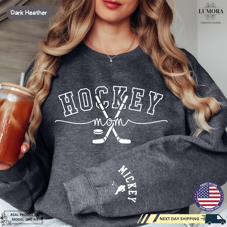 以下が含まれることがあります： ダークヘザーグレーのトレーナーに、白い文字で「HOCKEY MOM」と書いてあり、交差したホッケーのスティックとパックが描かれています。袖には「MICKEY」の文字がプリントされています。