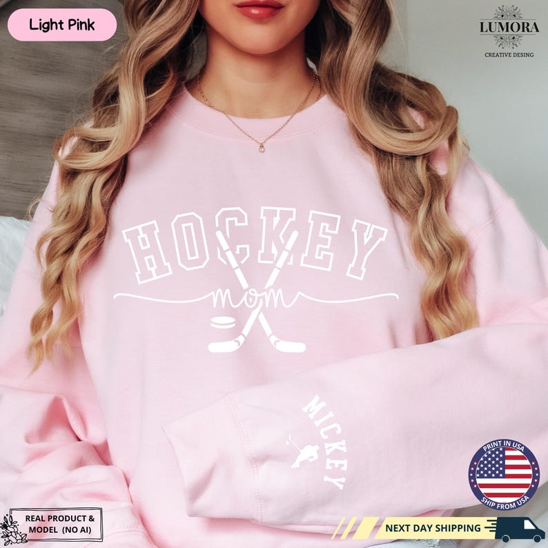 以下が含まれることがあります： 白地に「HOCKEY MOM」と書いてある薄いピンク色のスウェットシャツ。交差したホッケーのスティックとパックが描かれている。