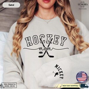 以下が含まれることがあります： 薄いグレーのトレーナーに、黒い文字で「HOCKEY MOM」と書いてあります。テキストには、交差した2本のホッケーのスティックとパックのグラフィックが添えられています。トレーナーの袖には「MICKEY」という文字が印刷されています。
