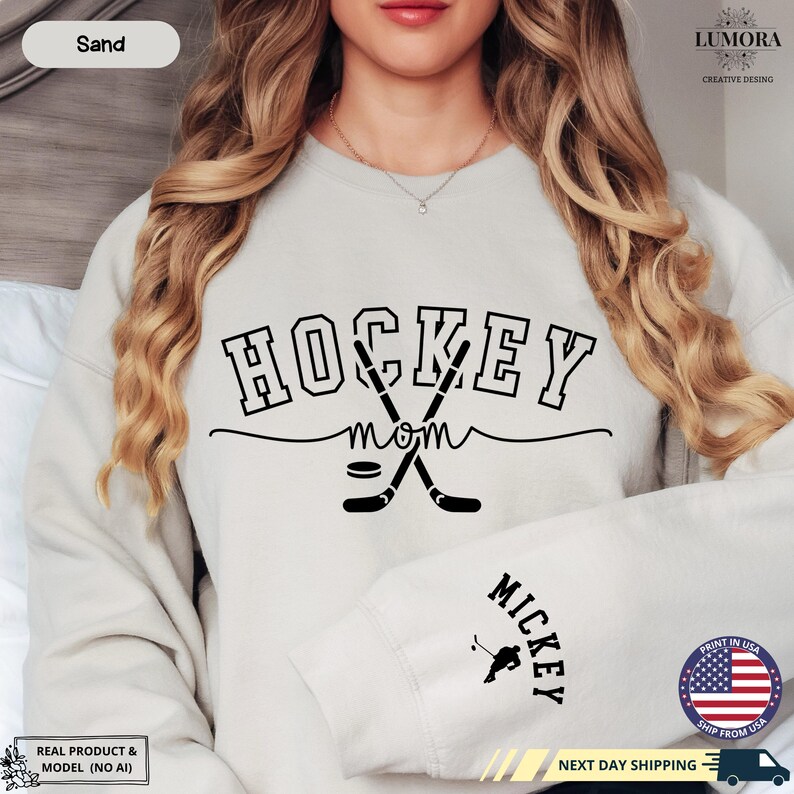 以下が含まれることがあります： 薄いグレーのトレーナーに、黒い文字で「HOCKEY MOM」と書いてあります。テキストには、交差した2本のホッケーのスティックとパックのグラフィックが添えられています。トレーナーの袖には「MICKEY」という文字が印刷されています。