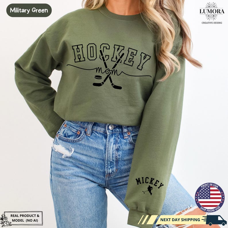 以下が含まれることがあります： 「HOCKEY mom」の文字とホッケーのスティックのグラフィックが描かれた緑色のスウェットシャツ。左袖には「MICKEY」の文字とホッケー選手のグラフィックが描かれている。