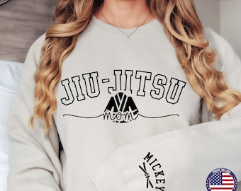 May include: A light gray sweatshirt with the text "JIU-JITSU" in black, with a stylized image of a person in a jiu-jitsu uniform and the word "mom" underneath. The text "MICKEY" is on the sleeve of the sweatshirt.