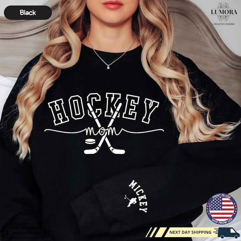 以下が含まれることがあります： 白い文字で「HOCKEY mom」と書いてある黒いスウェットシャツ。ホッケーのスティックとパックのグラフィックが描かれている。袖には「MICKEY」の文字が書かれている。