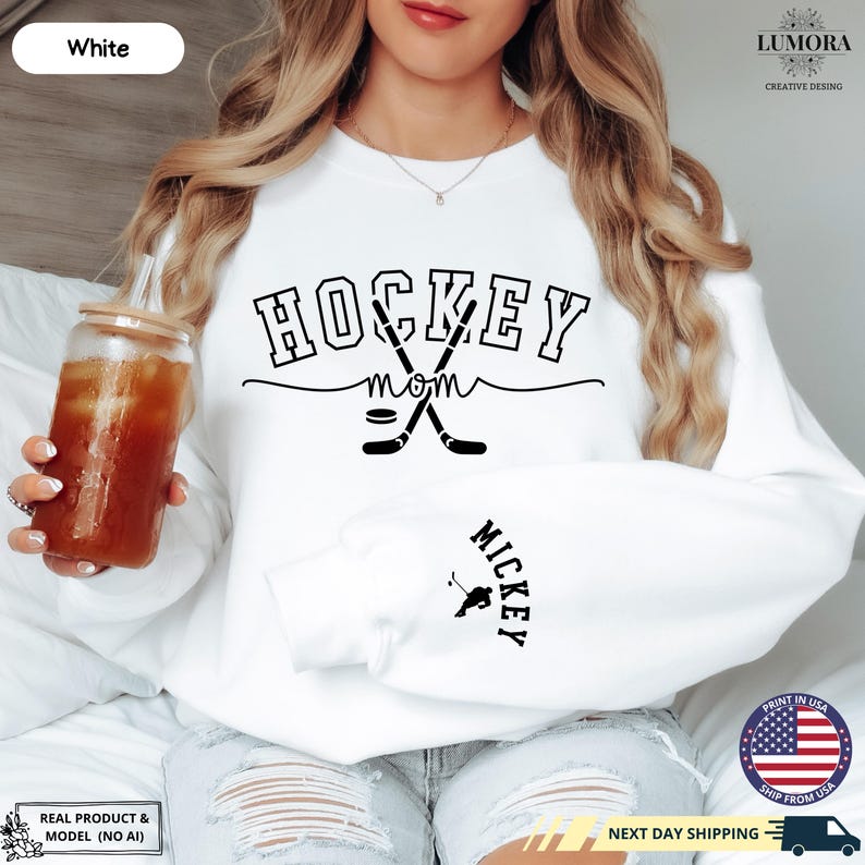以下が含まれることがあります： 黒い文字で「HOCKEY mom」と書いてある白いスウェットシャツ。交差したホッケーのスティックとパックが描かれている。テキストの下には、ホッケー選手の小さなグラフィックと「MICKEY」というテキストがある。