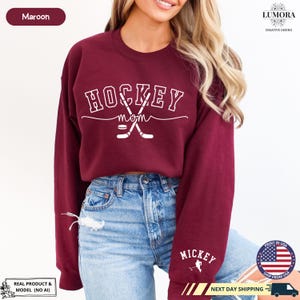 以下が含まれることがあります： 「HOCKEY mom」の文字と、交差した2本のホッケーのスティックとパックのグラフィックが描かれた、マルーンのトレーナー。トレーナーには袖に「MICKEY」の文字も書かれている。