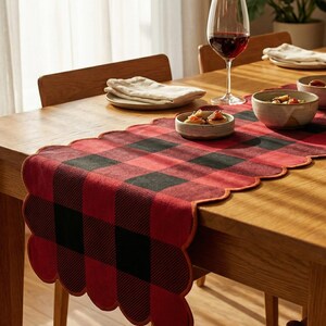 May include: A wooden dining table set for a meal, featuring a red and black buffalo check table runner with a scalloped edge. Wine glasses, small bowls, and folded napkins are arranged on the table.
