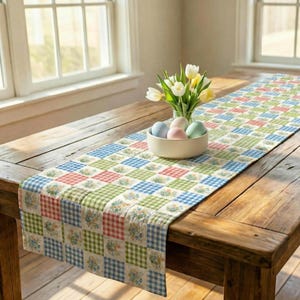 May include: A colorful table runner with a patchwork design in red, green, and blue squares, adorned with floral patterns. A wooden table holds a white bowl with pastel-colored eggs and a vase of white and yellow tulips.