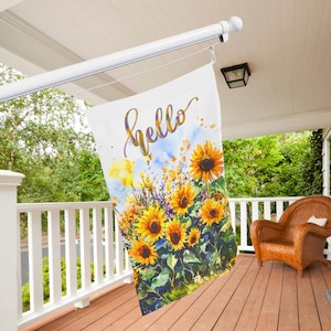 May include: A white garden flag with a floral design featuring sunflowers and the word "hello" in gold script. The flag is hanging on a white pole on a porch with a wooden floor and white railing.