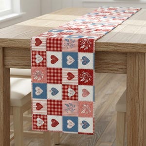 May include: A red, white, and blue patchwork table runner with heart and floral patterns. The runner is displayed on a wooden table, showcasing a country or farmhouse style. The runner's design includes gingham and solid squares.