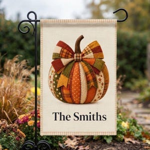 May include: A rectangular garden flag featuring a central pumpkin illustration composed of various autumn-colored fabric patterns and a large bow. The text The Smiths is printed in black below the pumpkin on a cream background with a checkered border.