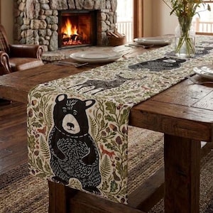 May include: A long rectangular table runner featuring a repeating pattern of black bears, deer, and foxes surrounded by green oak leaves, acorns, and ferns on a cream-colored fabric base, draped across a rustic wooden dining table.