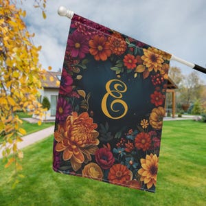 Puede incluir: Una bandera de jardín rectangular que presenta un centro azul marino oscuro con una gran letra E cursiva de color amarillo dorado. El borde está decorado con un vibrante arreglo de flores de colores otoñales, incluyendo crisantemos naranjas, rojos y amarillos.
