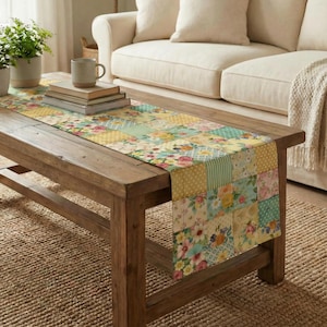 May include: A patchwork table runner with a floral pattern in pastel colors of yellow, pink, and blue. The runner is placed on a rustic wooden coffee table, with a beige sofa in the background. A stack of books and a mug are on the table.