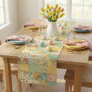 Op de afbeelding: Een houten eettafel gedekt voor een maaltijd, met een bloemen patchwork tafelloper. De tafel is gedekt met borden, servetten en goudkleurig bestek. Een witte vaas bevat gele en roze bloemen, en decoratieve paaseieren zijn verspreid.