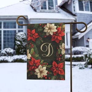 Peut inclure: Un drapeau de jardin avec un motif festif de poinsettias rouges et crème, de feuillage vert et de baies rouges. Le drapeau a un fond sombre avec une grande lettre "D" au centre. Le drapeau est affiché sur un support en métal.
