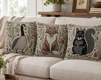 May include: Three square throw pillows featuring woven illustrations of a Canada goose, a red fox, and a black squirrel. Each pillow is decorated with detailed botanical patterns including ferns, acorns, and small woodland creatures on a beige background.