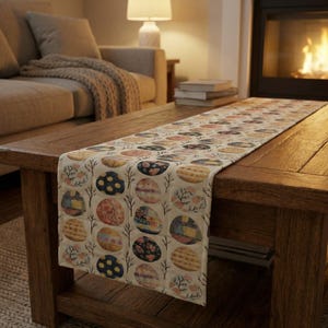 May include: A beige Easter-themed table runner with a repeating pattern of colorful, decorated Easter eggs. The eggs feature various designs, including floral, geometric, and abstract patterns. The runner is displayed on a wooden coffee table.