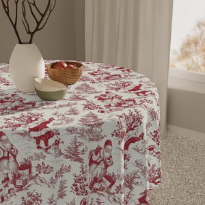 Peut inclure: Une nappe ronde avec un motif de Noël rouge et blanc. Le motif représente des figures du Père Noël, des arbres et des scènes d'hiver. La nappe est drapée sur une table ronde, avec un vase, des bols et un panier de pommes.