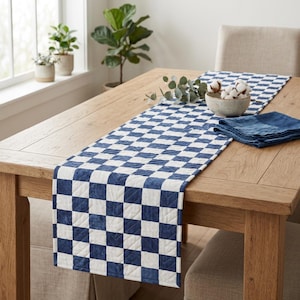 May include: A blue and white chequered table runner on a light brown wooden table. A matching blue napkin and a bowl of cotton balls are also on the table. The runner is quilted and adds a decorative touch to the dining area.
