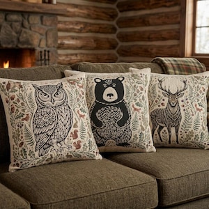 May include: Three square throw pillows featuring detailed illustrations of a black owl, a black bear, and a brown deer. Each pillow is decorated with a botanical pattern of green leaves, acorns, and small woodland animals on a light beige fabric.