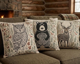 May include: Three square throw pillows featuring detailed illustrations of a black owl, a black bear, and a brown deer. Each pillow is decorated with a botanical pattern of green leaves, acorns, and small woodland animals on a light beige fabric.