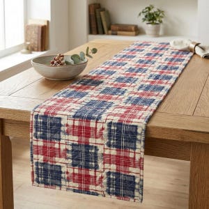 May include: A rectangular table runner with a quilted pattern in red, white, and navy blue. The design features a plaid-like arrangement of squares and rectangles. The runner is placed on a wooden table.