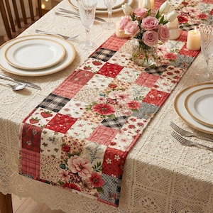 Pode incluir: Um caminho de mesa acolchoado com um design de patchwork com quadrados vermelhos, brancos e pretos. Os quadrados exibem padrões florais, xadrez e morangos. O caminho de mesa é colocado sobre uma toalha de mesa de renda branca, com pratos, talheres e um centro de mesa floral.
