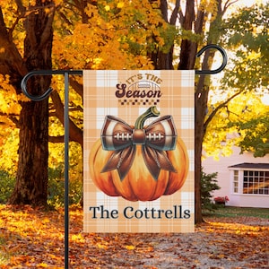 May include: A garden flag with a pumpkin design and a football-themed bow. The flag displays the text "It's the Season" and "The Cottrells" on a plaid pattern. The flag is on a black metal stand, set against a backdrop of autumn leaves.