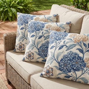 May include: Three square throw pillows featuring a botanical pattern of blue and beige hydrangea flowers and stylized floral motifs on a cream background. The pillows are arranged on a light beige outdoor sofa with a woven wicker frame.