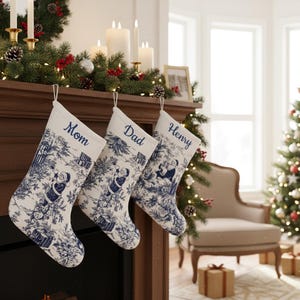 Puede incluir: Tres calcetines navideños blancos con diseños azules cuelgan sobre una chimenea. Cada calcetín tiene un nombre bordado: Mamá, Papá e Henry. La chimenea está decorada con vegetación, velas y una foto enmarcada.