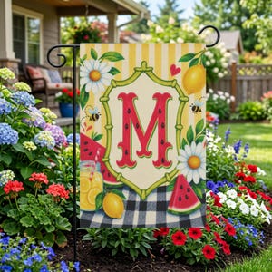 Op de afbeelding: Een decoratieve tuinvaandel met een grote rode letter "M" met een watermeloenontwerp. De vlag heeft een geel en wit gestreepte achtergrond, met illustraties van citroenen, madeliefjes, bijen en watermeloenschijfjes. De vlag wordt in een tuin getoond.