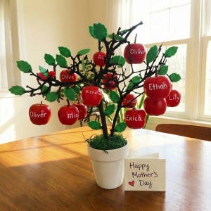May include: A decorative tree with red apple ornaments, each bearing a name in white script. The tree sits in a white pot on a wooden table, with a card that reads "Happy Mother's Day". Green leaves and moss add to the design.