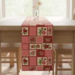 May include: A rectangular table runner with a patchwork design featuring red roses on a cream background, framed by red squares and stripes. The runner is placed on a wooden table, with a vase of flowers in the background.