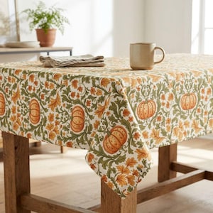 May include: A rectangular tablecloth featuring a repeating pattern of orange pumpkins and green foliage on a cream background. A folded beige napkin and a plain, light-colored ceramic mug are placed on the surface of the table.