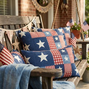 May include: Two rectangular throw pillows featuring a patchwork design of navy blue fabric with white stars and red and white gingham and striped patterns. The pillows are arranged on a dark wooden bench decorated with patriotic bunting and flags.