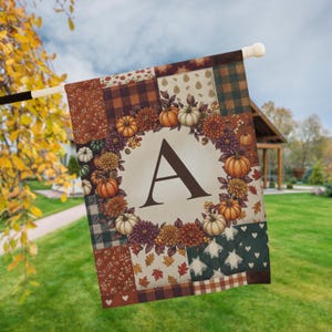 Peut inclure: Un drapeau de jardin rectangulaire présentant un motif patchwork avec des thèmes automnaux, incluant des carreaux, des fleurs et des cœurs. Un cercle central crème affiche une grande lettre A marron entourée d'une couronne de citrouilles.