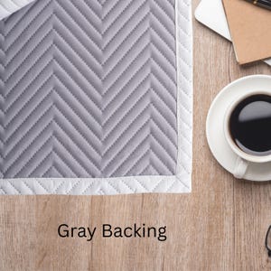 Op de afbeelding: Een grijze gewatteerde stof met een chevronpatroon en witte rand. De tekst "Gray Backing" is onderaan afgedrukt. Een witte koffiekop met zwarte koffie staat op een houten oppervlak.