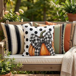 May include: Three square throw pillows arranged on a wooden bench. One pillow features black and cream vertical stripes. The center pillow is white with black polka dots and an orange pumpkin graphic tied with a black and white buffalo check bow.