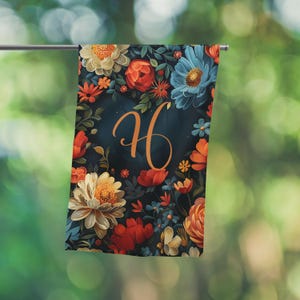 Puede incluir: Una toalla con estampado floral con un fondo azul oscuro y la letra "H" en escritura naranja. La toalla presenta una variedad de flores en tonos de rojo, naranja, azul y crema.