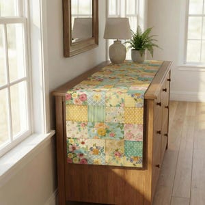 Op de afbeelding: Een bloemen patchwork tafelloper in tinten geel, roze en blauw. De loper is geplaatst op een houten dressoir. Het ontwerp heeft vierkanten met bloempatronen, stippen en strepen, wat een vintage esthetiek cre&euml;ert.