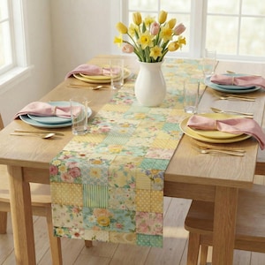 Op de afbeelding: Een houten eettafel gedekt voor een maaltijd, met een bloemen patchwork tafelloper in pastelkleuren. Gele en roze borden met roze servetten zijn voor elke gast gedekt. Een witte vaas bevat gele en roze bloemen.